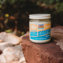 Load image into Gallery viewer, Lake Geneva soy candle in a 9 oz glass jar, from the Wanderlust Wisconsin Collection by Angela Rose Studios, shown outdoors on a rock with a blurred natural, outdoor-naturesque setting in the background.