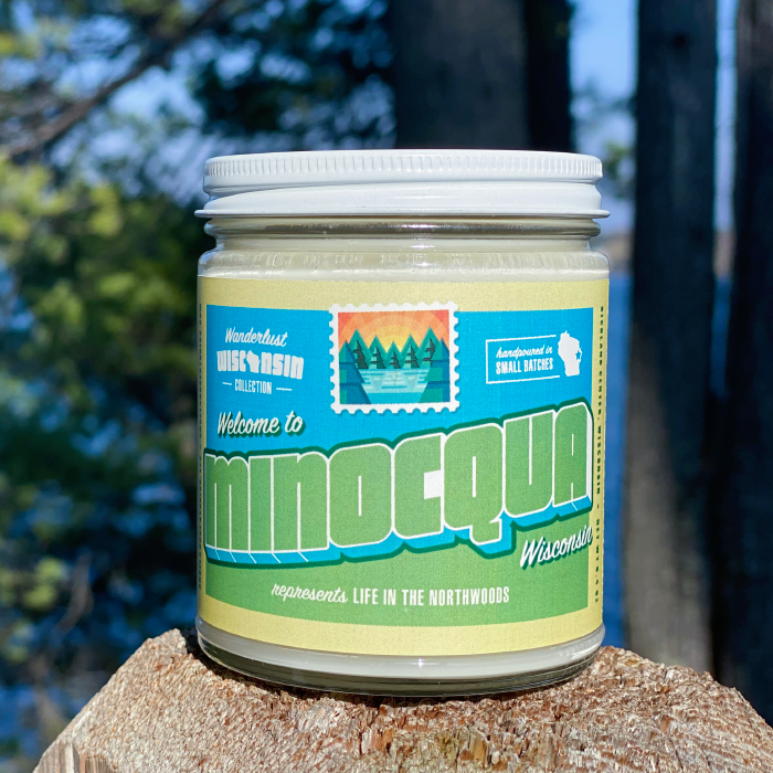 Minocqua soy candle in a 9 oz glass jar, from the Wanderlust Wisconsin Collection by Angela Rose Studios, shown outdoors on a tree stump with a blurred natural, outdoor-naturesque forest background.