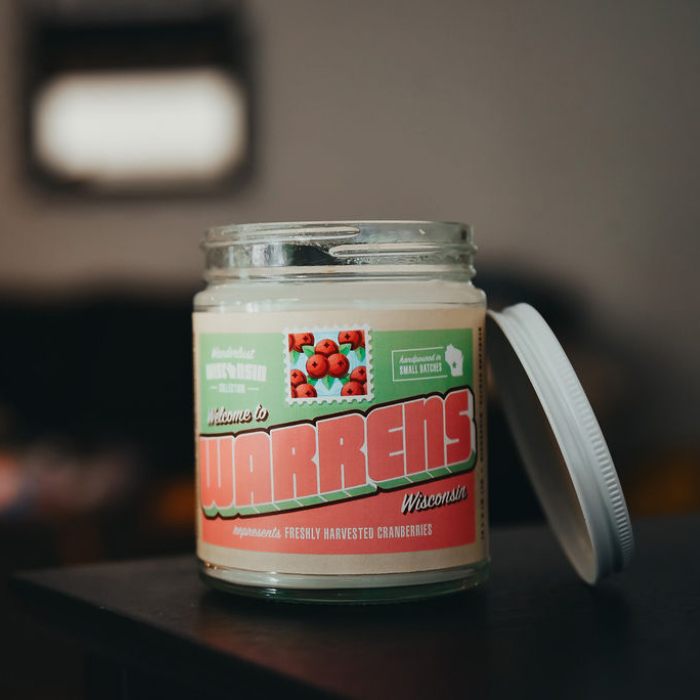 Warrens soy candle in a 9 oz glass jar, from the Wanderlust Wisconsin Collection by Angela Rose Studios, shown indoors on a dark wooden surface with a softly blurred, warm-toned background and the candle lid resting off to the side.